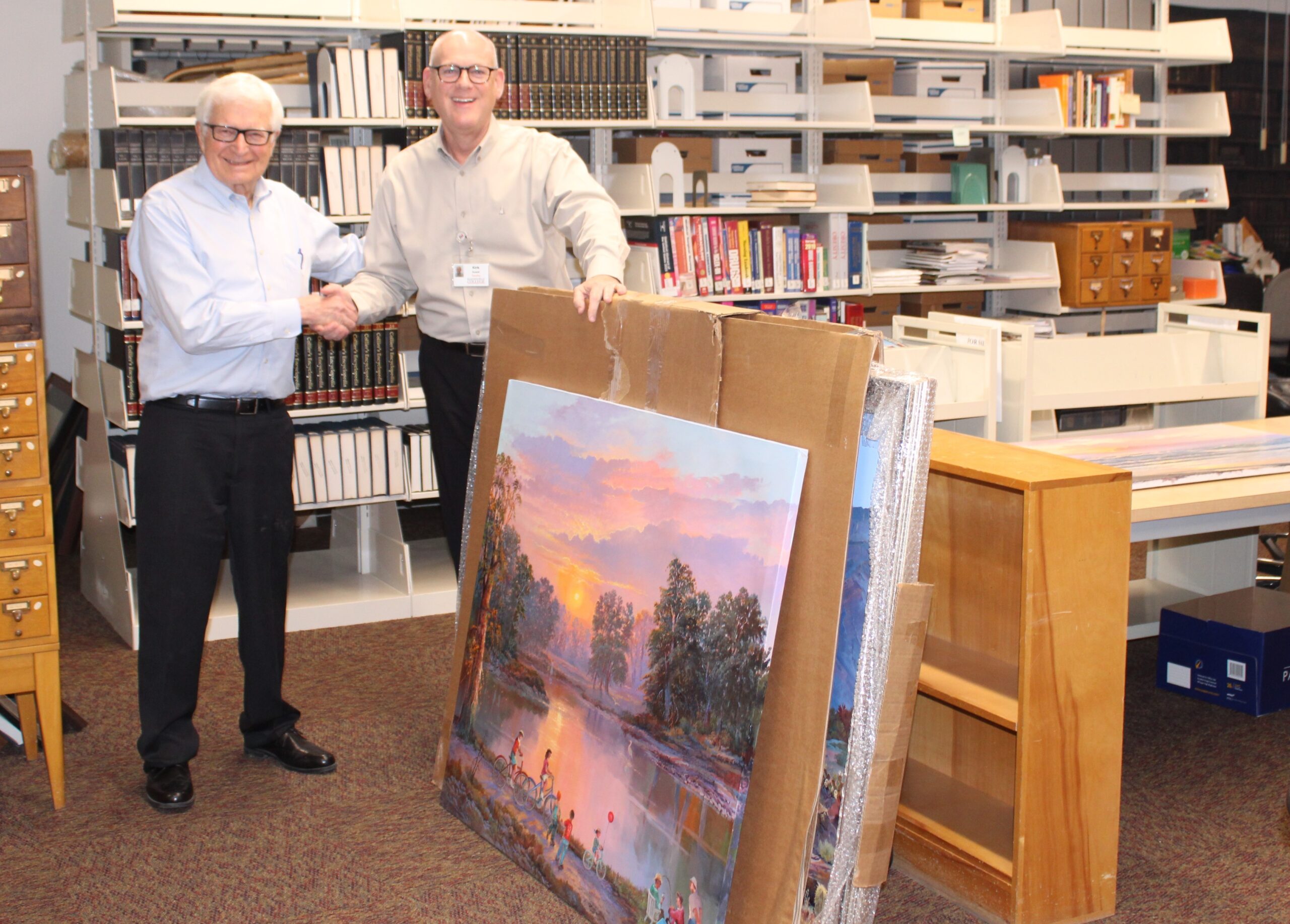 Ken Byrum shaking Kirk Russell's hand standing next to one of Beverly Carrick's paintings. 