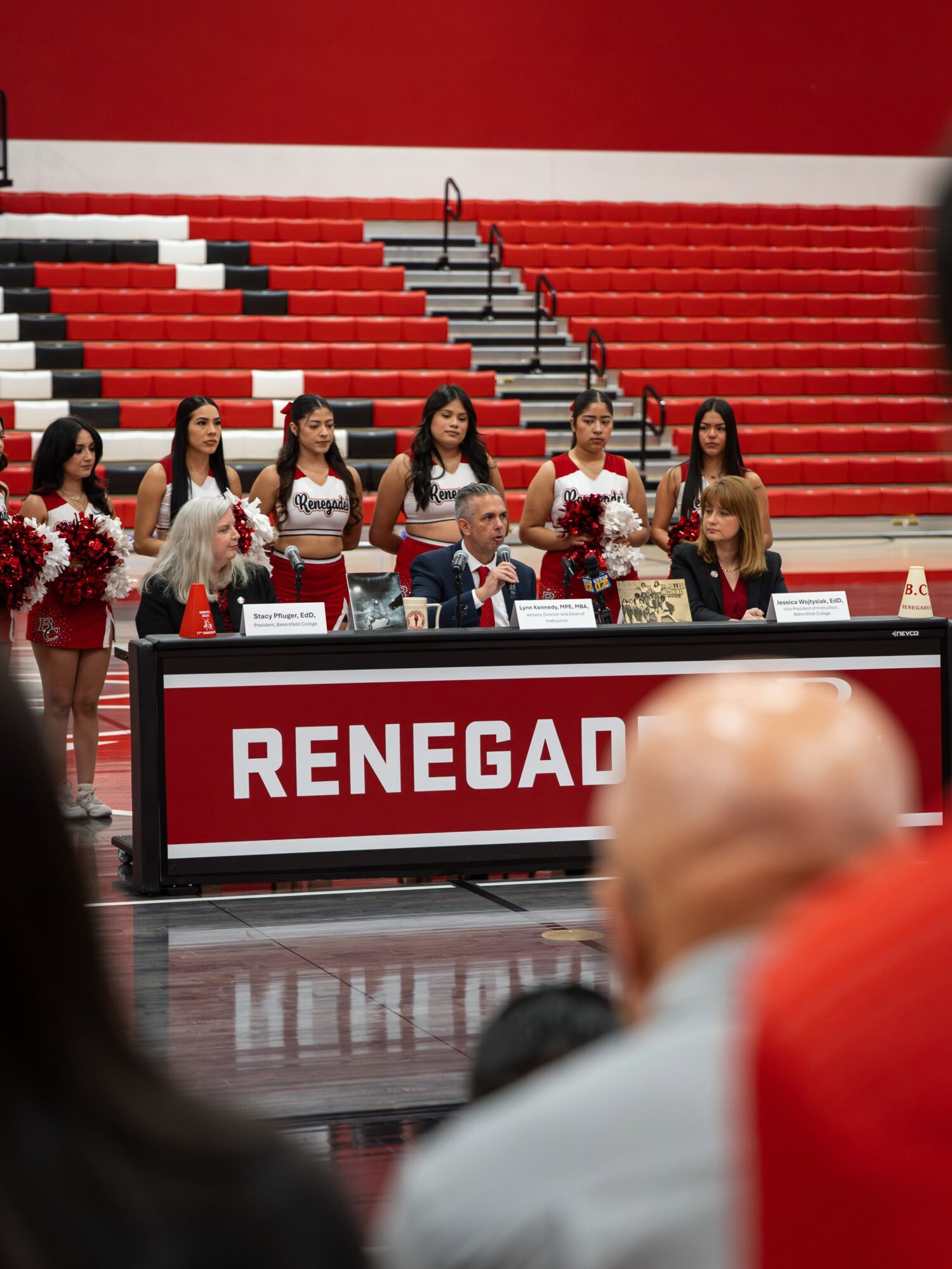 BC leaders speak at a gymnasium panel as cheerleaders stand behind them and spectators look on.